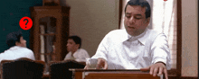 a man in a white shirt is sitting at a desk with a red question mark in the corner