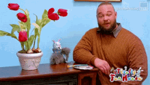 a man in a brown sweater sits in front of a table with a potted plant and a stuffed animal on it .