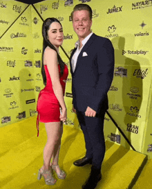 a woman in a red dress stands next to a man on a yellow carpet that says totalplay