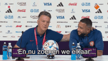 two men are sitting at a table with a soccer ball in front of a wall with logos for qatar airways and visa