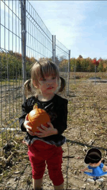 a little girl is holding a pumpkin in front of a wire fence