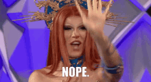 a drag queen with red hair and a crown on her head is waving her hand and saying nope .