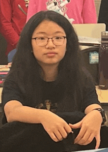 a girl wearing glasses and a black shirt is sitting at a desk .