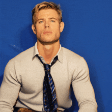 a man wearing a tie and a sweater is sitting in front of a blue background