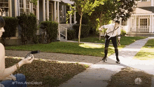 a man is rollerblading down a sidewalk in front of a house while a woman holds a remote control .