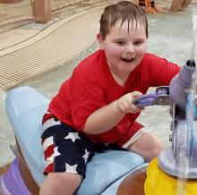 a young boy in a red shirt is sitting on a toy boat in a pool