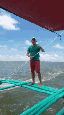 a man in a green shirt and red shorts is standing on a boat in the ocean giving a thumbs up