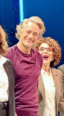 a man in a purple shirt smiles while standing next to a woman in glasses