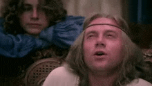 a man with long hair and a headband is sitting on a couch with another man .