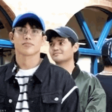 a man wearing glasses and a baseball cap is standing next to another man wearing a jacket .