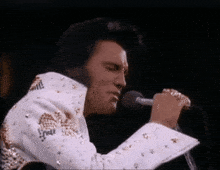 elvis presley is singing into a microphone while wearing a white and gold suit .