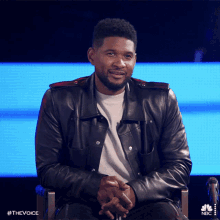 a man in a leather jacket is sitting in a chair with his hands folded in front of a nbc screen