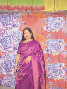 a woman in a purple saree is standing in front of a colorful floral curtain