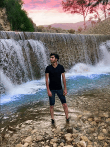 a man stands in front of a waterfall with a pink sky in the background