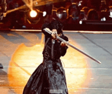 a woman in a black dress is holding a sword on a stage in front of a crowd .