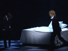a man in a suit stands in front of a bed with white sheets