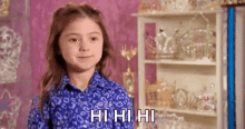 a little girl in a blue shirt is standing in front of a shelf full of crowns and tiaras .