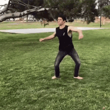 a man in a black tank top and jeans is standing in the grass in front of a tree .