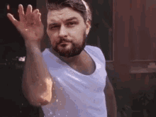 a man with a beard is wearing a white tank top and is waving his hand .