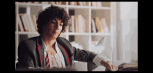 a woman with curly hair is sitting at a desk in front of a bookshelf and looking at her watch .