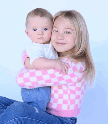 a girl in a pink and white checkered sweater holds a baby