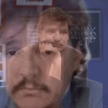 a man with a mustache is sitting down with his hand on his chin and looking at the camera .