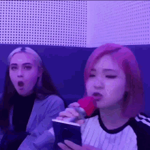 a girl with pink hair is singing into a microphone while another girl looks surprised .