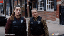 two female police officers standing next to each other in front of a brick building with nbc written on the bottom