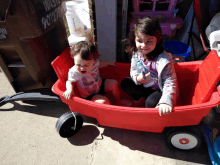 two little girls are sitting in a red wagon in front of a waste container