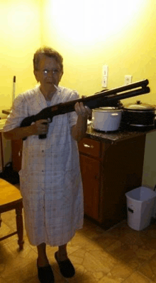 an elderly woman is holding a shotgun in a kitchen and smoking a cigarette