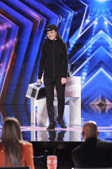 a woman in a black jacket and hat stands on a stage holding a box and smiling