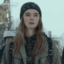 a girl wearing a beanie and a jacket is standing in the snow with jurassic world dominion written above her