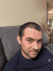 a man with a beard is sitting on a couch wearing a blue shirt with an embroidered bird on it