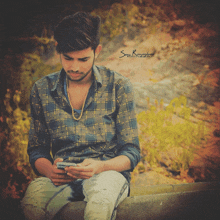 a young man in a plaid shirt sits on a ledge looking at his cell phone with a photo by sony rameshwar