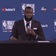 a man in a suit and tie is sitting at a table with a microphone in front of a nba finals sign