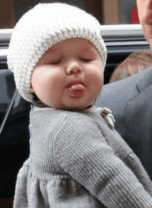 a baby wearing a white hat and a gray sweater is sticking out his tongue .