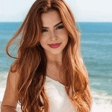 a woman with long red hair is standing on the beach and smiling at the camera .