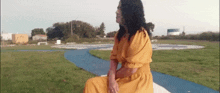 a woman in a yellow dress is sitting on a bench in front of a grassy field .