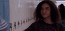 a young woman with curly hair is standing in front of a row of lockers in a school hallway .