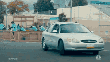 a silver ford taurus is driving down a street in front of a group of people working in a garden .