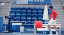 a man in a red shirt and white shorts is holding a tennis racquet in a tokyo 2020 stadium .