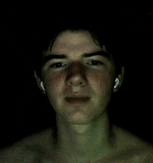 a shirtless young man wearing ear buds looks at the camera in the dark