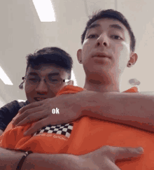 a man wearing an orange shirt with a checkered pattern is being hugged by another man who says ok