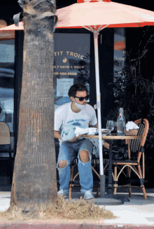 a man wearing a mask sits at a table under an umbrella in front of a restaurant called tit trois