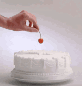 a hand holds a cherry over a white cake