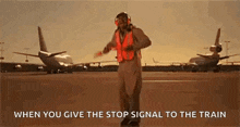 a man in an orange vest is standing on an airport runway giving a stop signal to an airplane .