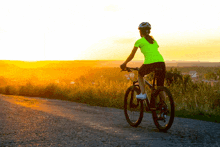 a woman wearing a helmet is riding a bike down a dirt road at sunset