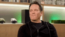 a man wearing a black hoodie is sitting in front of a bookshelf .