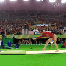 a woman in a red leotard is doing a trick on a mat with the olympic rings in the background