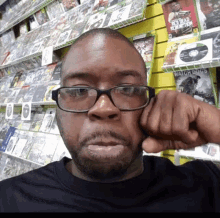 a man wearing glasses holds his fist to his face in front of a wall of video games including red dead redemption and watchdogs